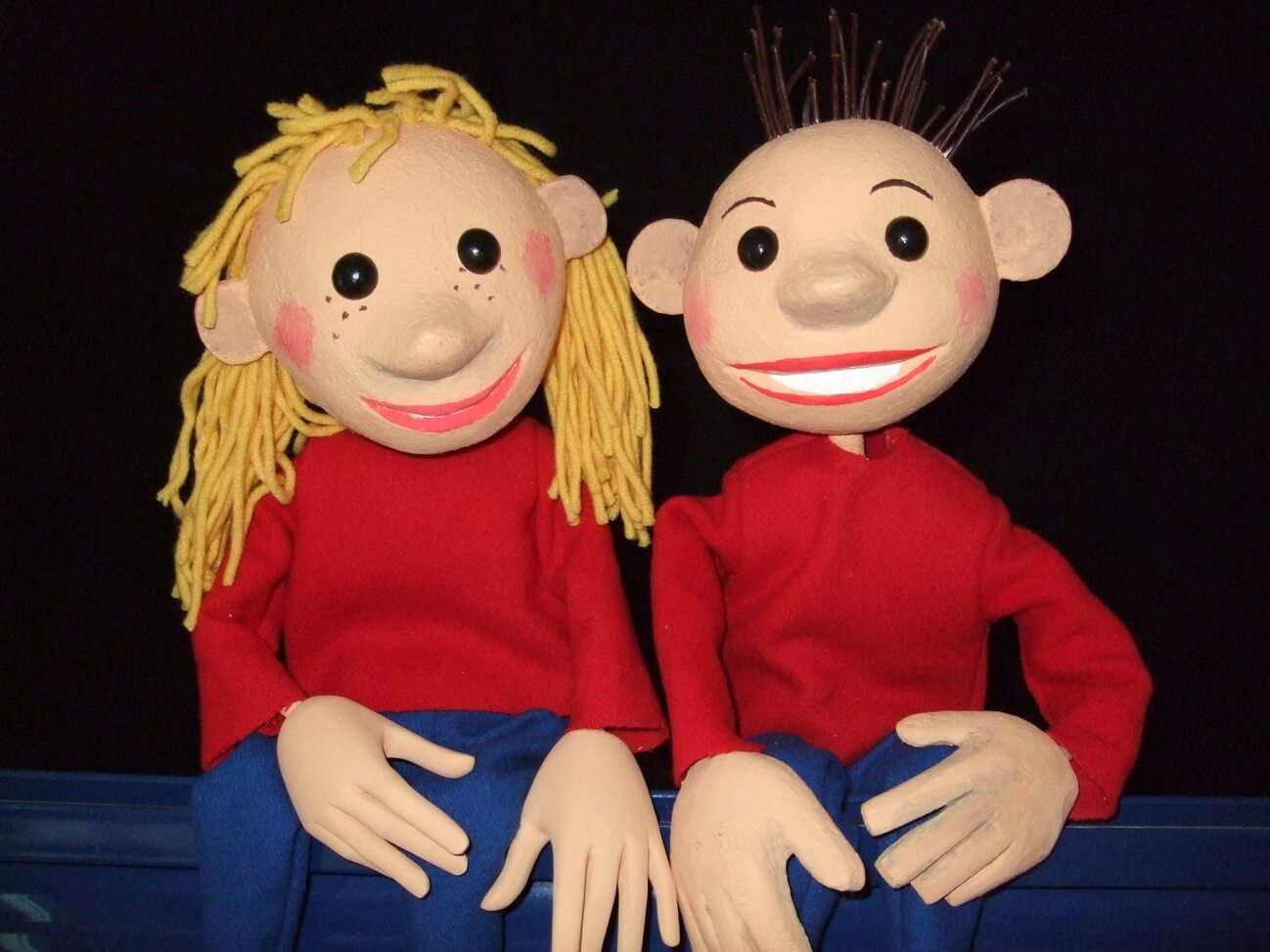 Foto: Foto der beiden Tischfiguren Flummi (Mädchen) und Flo (Junge).Beide haben rote Pullover und blaue Hosen an.